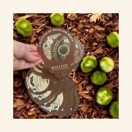 Wild Life Poo Clues flip-card guide held outdoors over wood chips and green seed pods, showing the round scat identification cards being used during nature exploration.